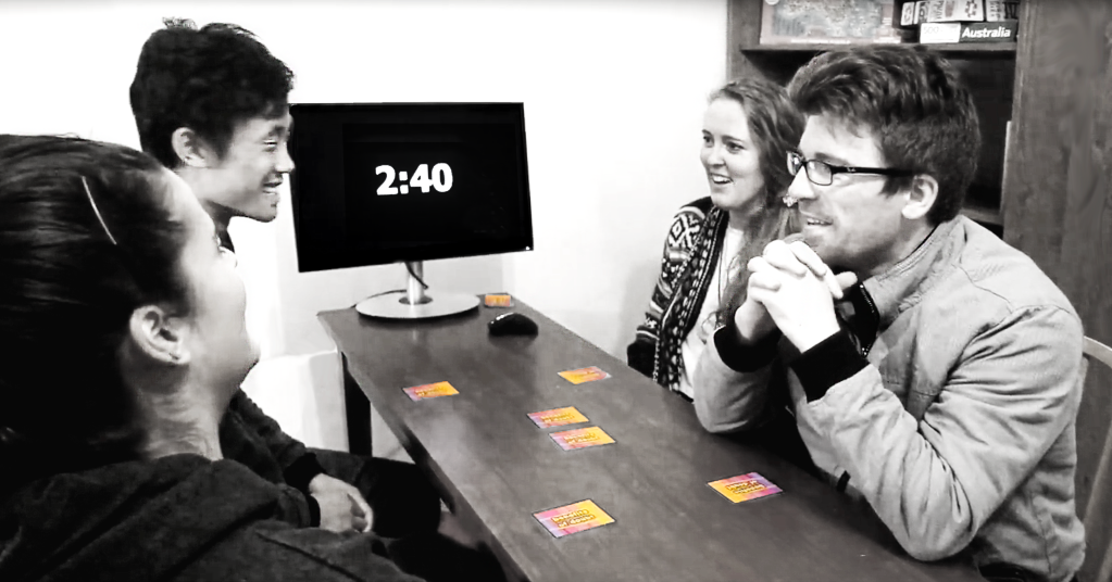 Four players sitting at a wooden table playing the game with 2 minutes and 40 seconds left on the clock. The character cards are face-down on the table and players are smiling at eachother during the discussion.